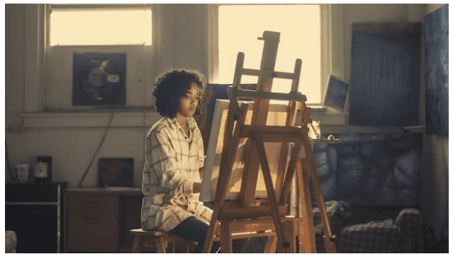 A woman sits on a stool, focused on her canvas at an easel, surrounded by art supplies and a creative atmosphere.